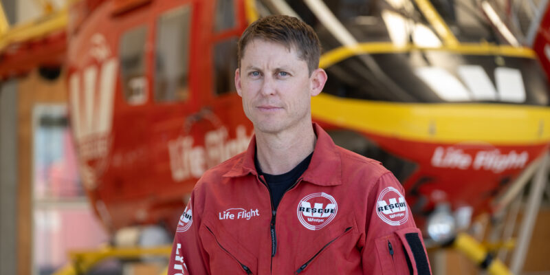 Caucasian, brunette man in helicopter flight suit standing in front of the Westpac Rescue Helicopter
